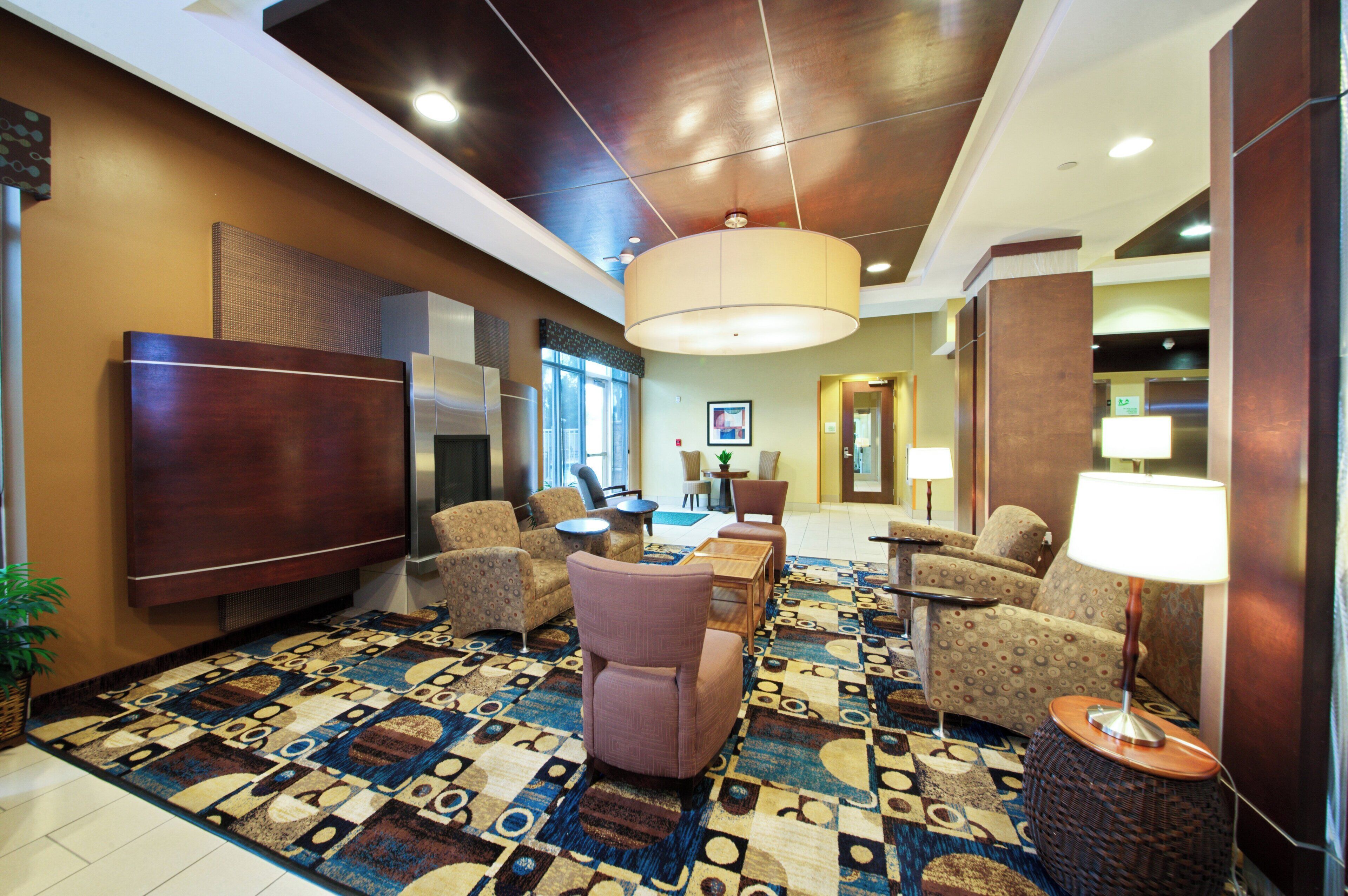 A hotel lobby with a large flat-screen TV, comfortable seating, a patterned carpet, and a modern ceiling design.