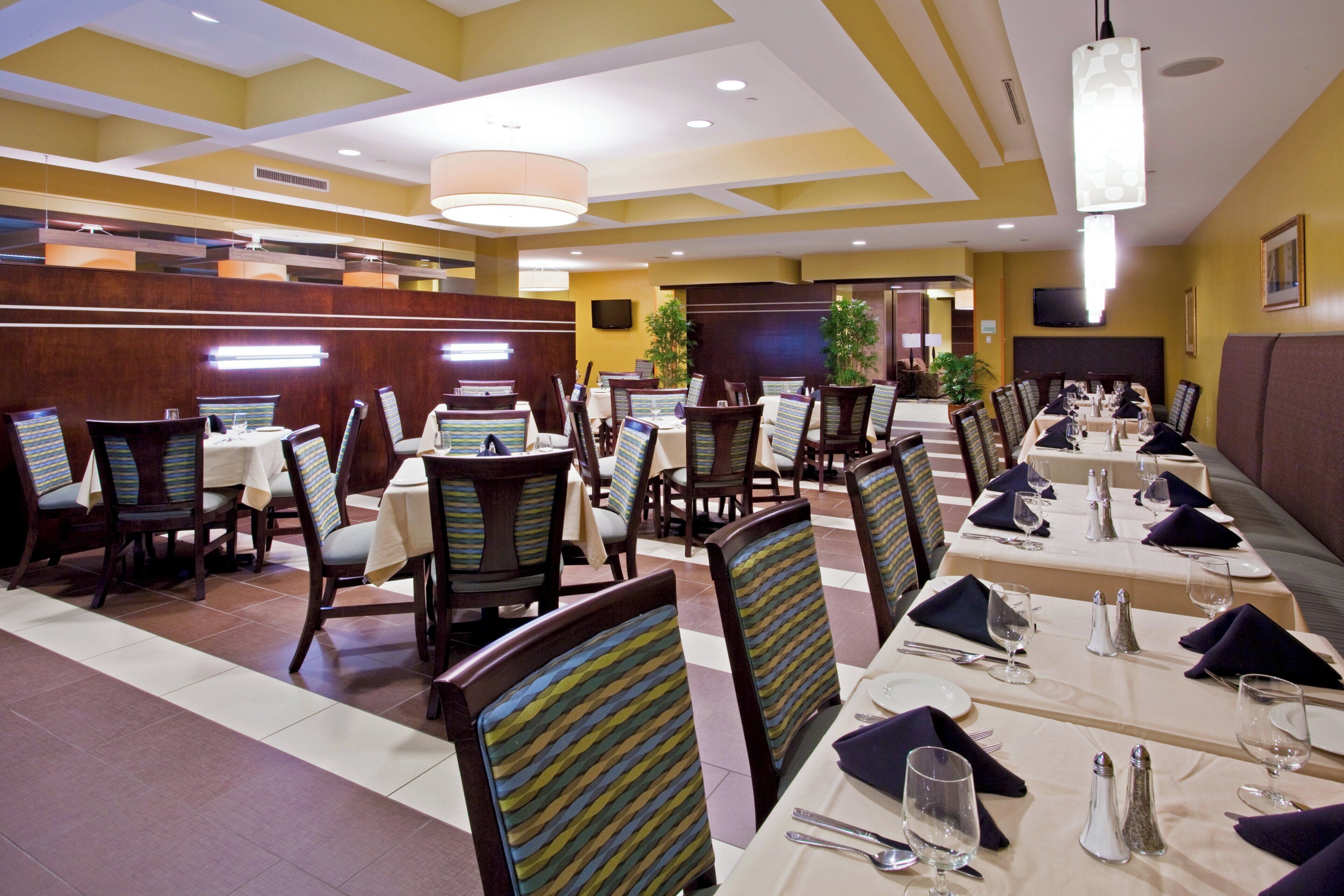 A hotel dining area with multiple tables and chairs, a television mounted on the wall, and a ceiling with recessed lighting.