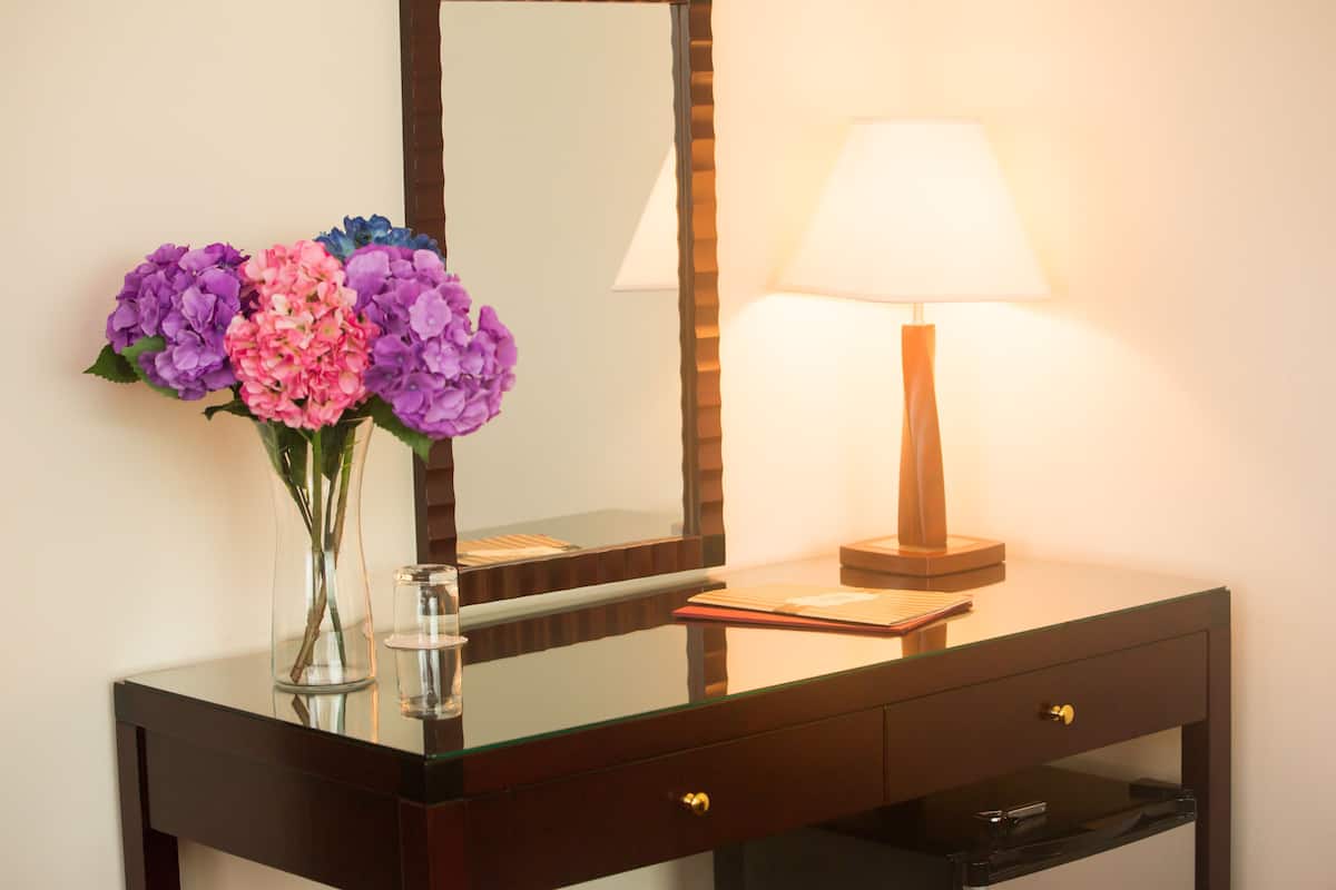A hotel room with a glass-top vanity, a mirror, a bedside lamp, and a vase with flowers.