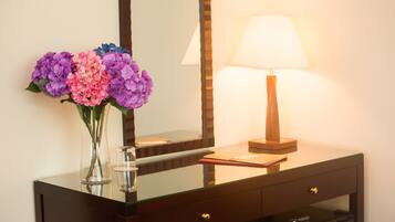 Een hotelkamer met een wastafel met glazen blad, een spiegel, een bedlamp en een vaas met bloemen.