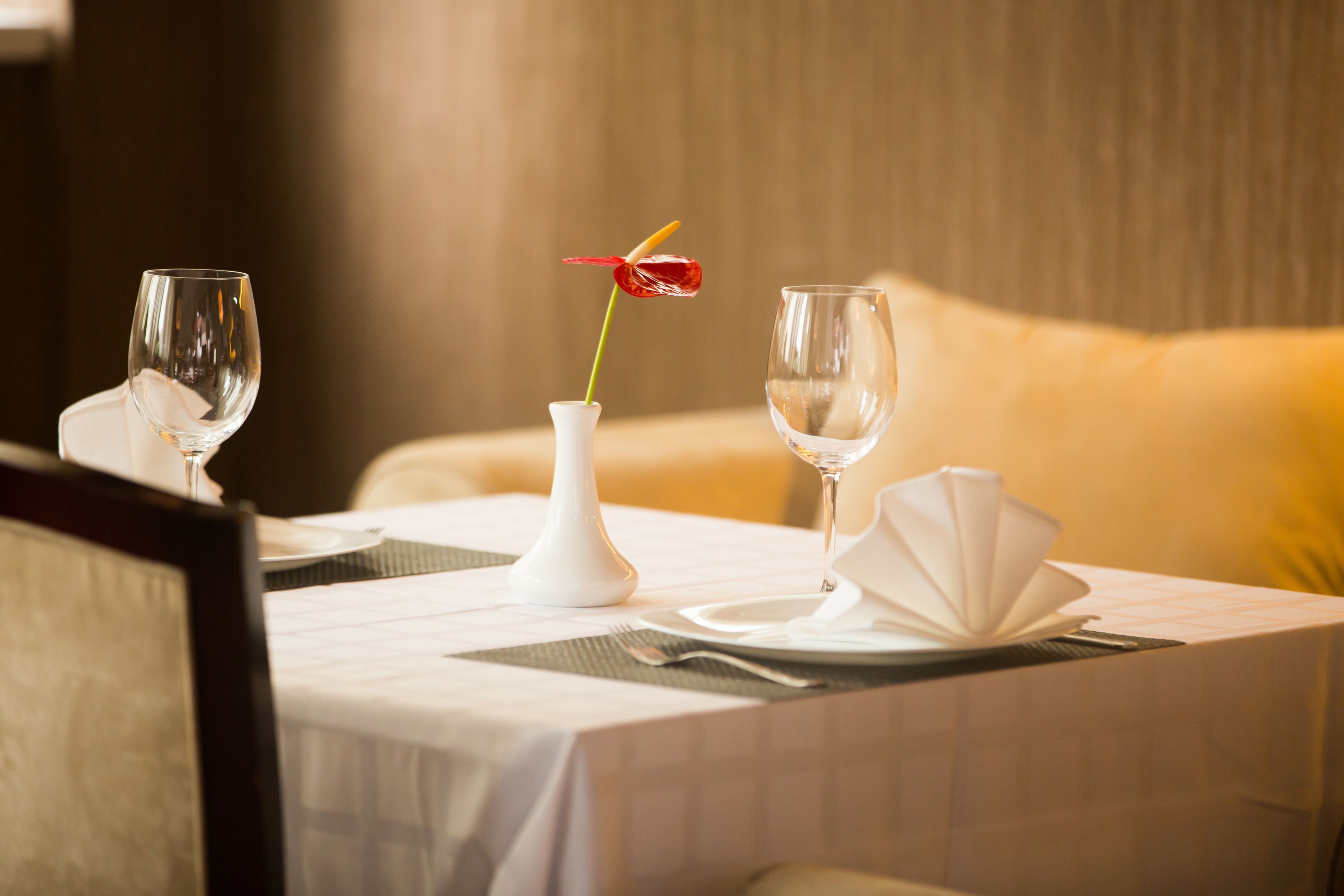 Una mesa de comedor bien dispuesta con dos copas de vino, un jarrón blanco con una flor roja y una servilleta doblada.