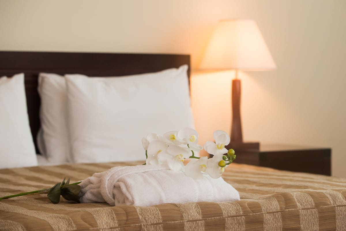 A neatly made bed with white pillows, a folded white towel, and a bouquet of white flowers.