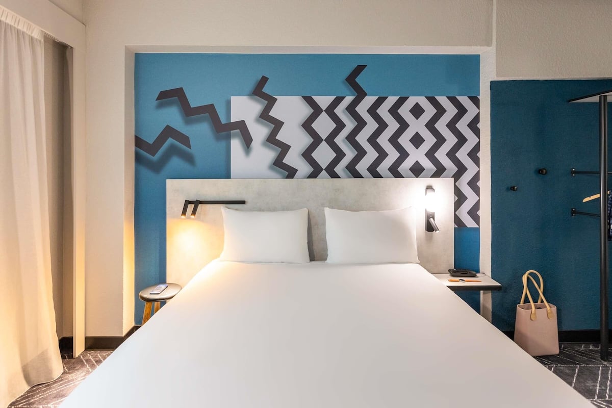 Una camera d'albergo con un letto grande, una testiera con un motivo geometrico sul muro, un comodino con una lampada e una porta con portasciugamani.