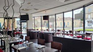 A modern restaurant with tables set for dining, large windows offering a view of the street, and a black and red color scheme. - ibis Styles Bordeaux Meriadeck (Bordeaux)