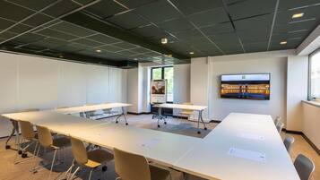 Un salón de conferencias con mesas blancas, sillas, una pantalla grande y un ventanal.