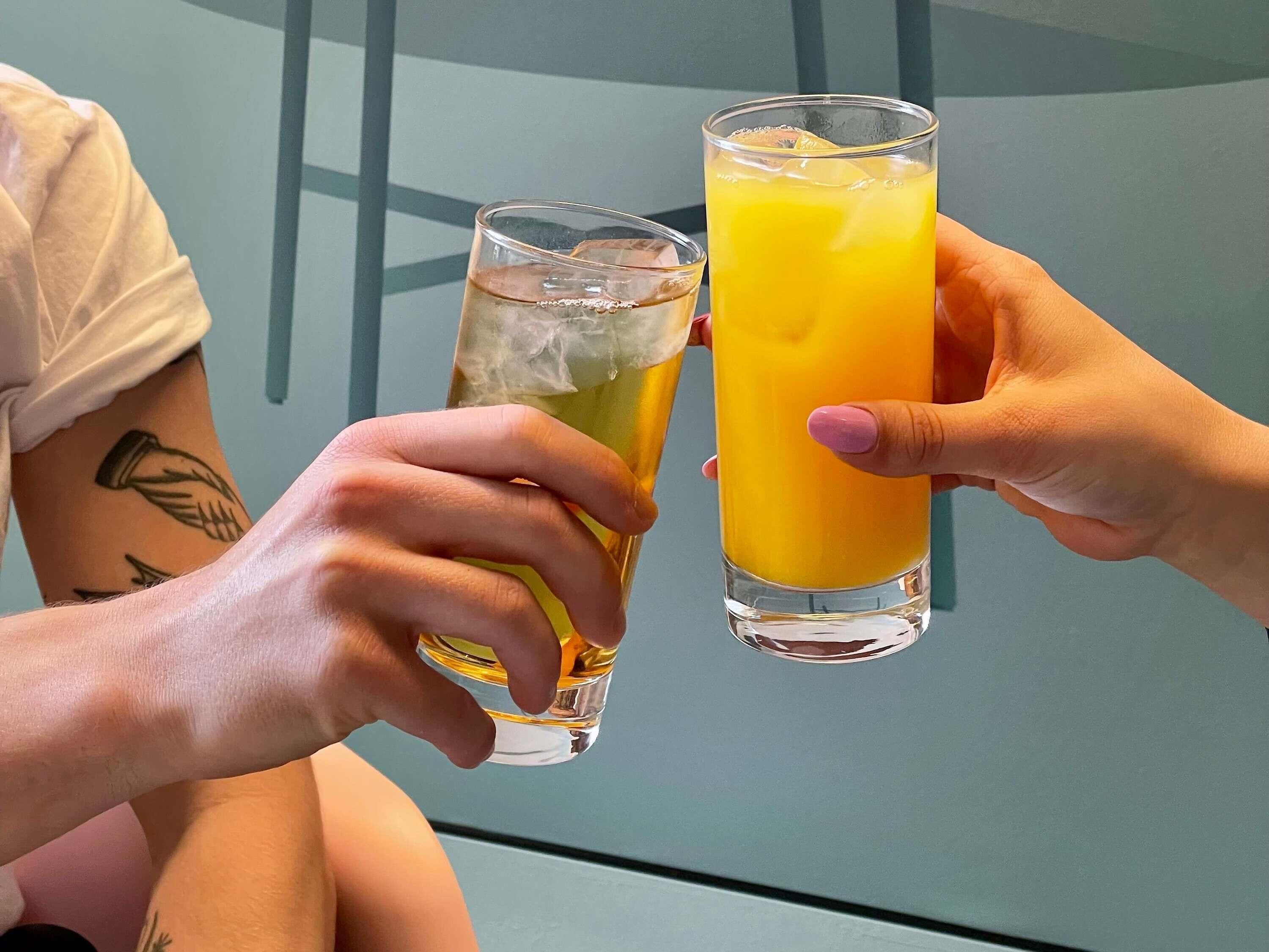 Duas pessoas segurando copos com bebidas, um com líquido transparente e outro com suco de laranja.