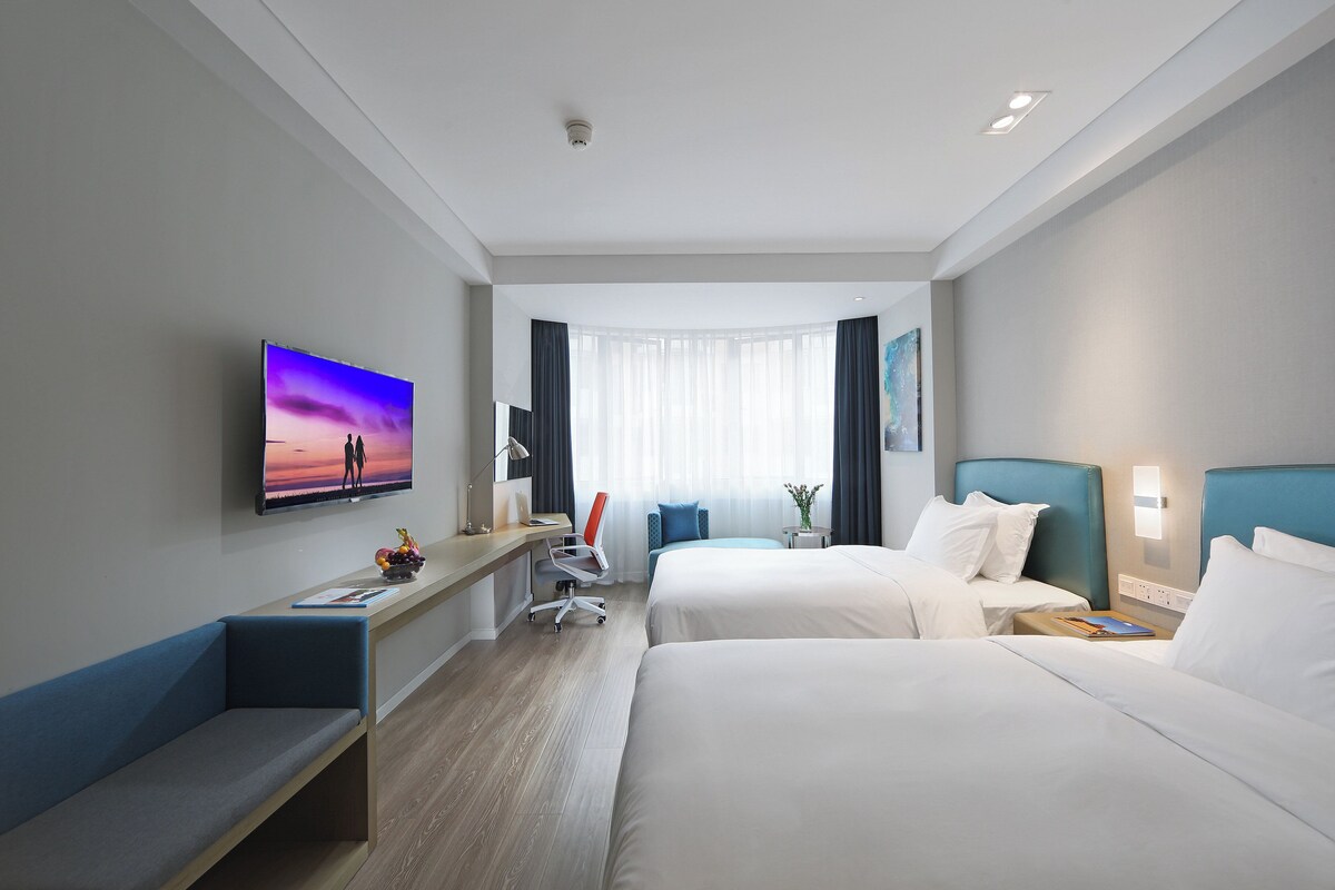A hotel room with two beds, a desk with a chair, a TV, and a window with curtains.