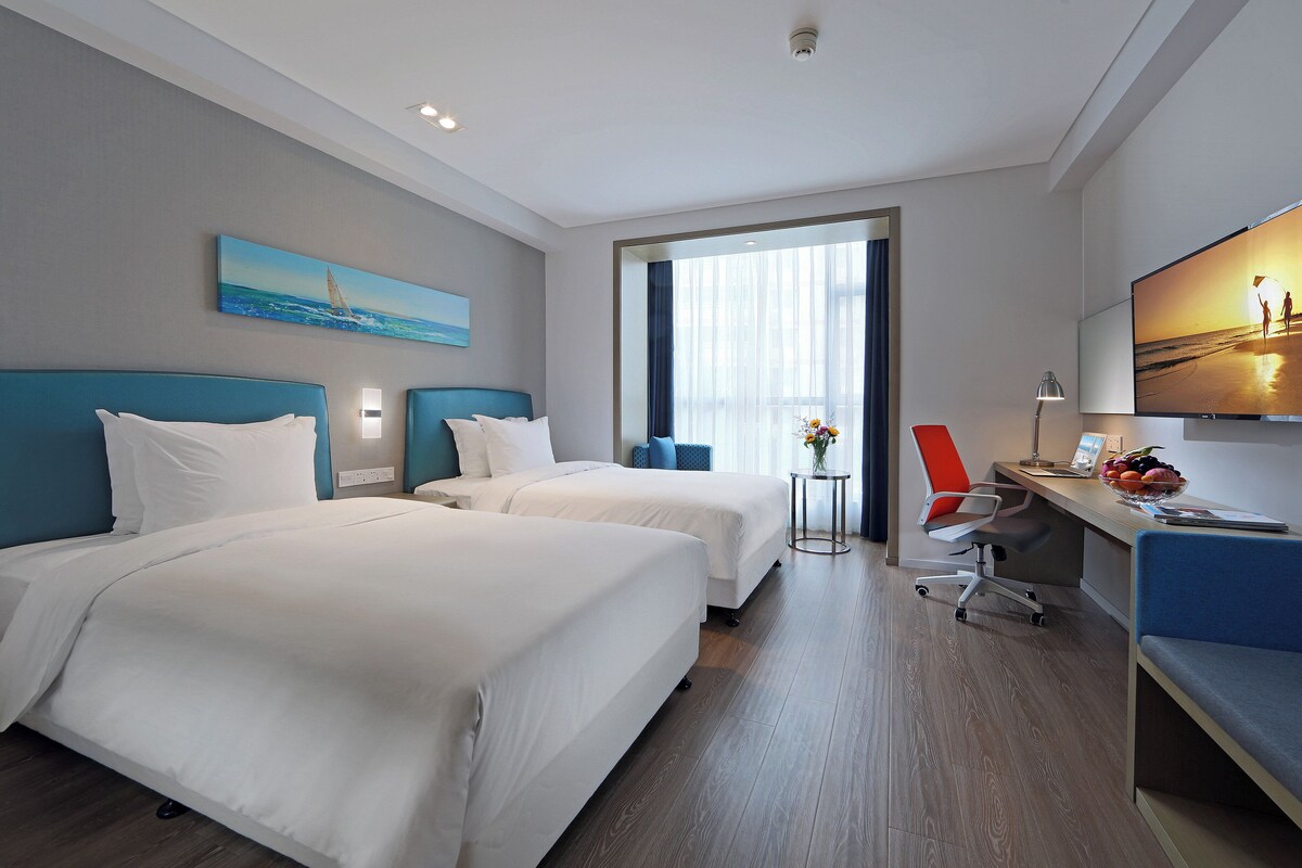 A hotel room with two beds, a desk with a chair, a TV, and a window with curtains.