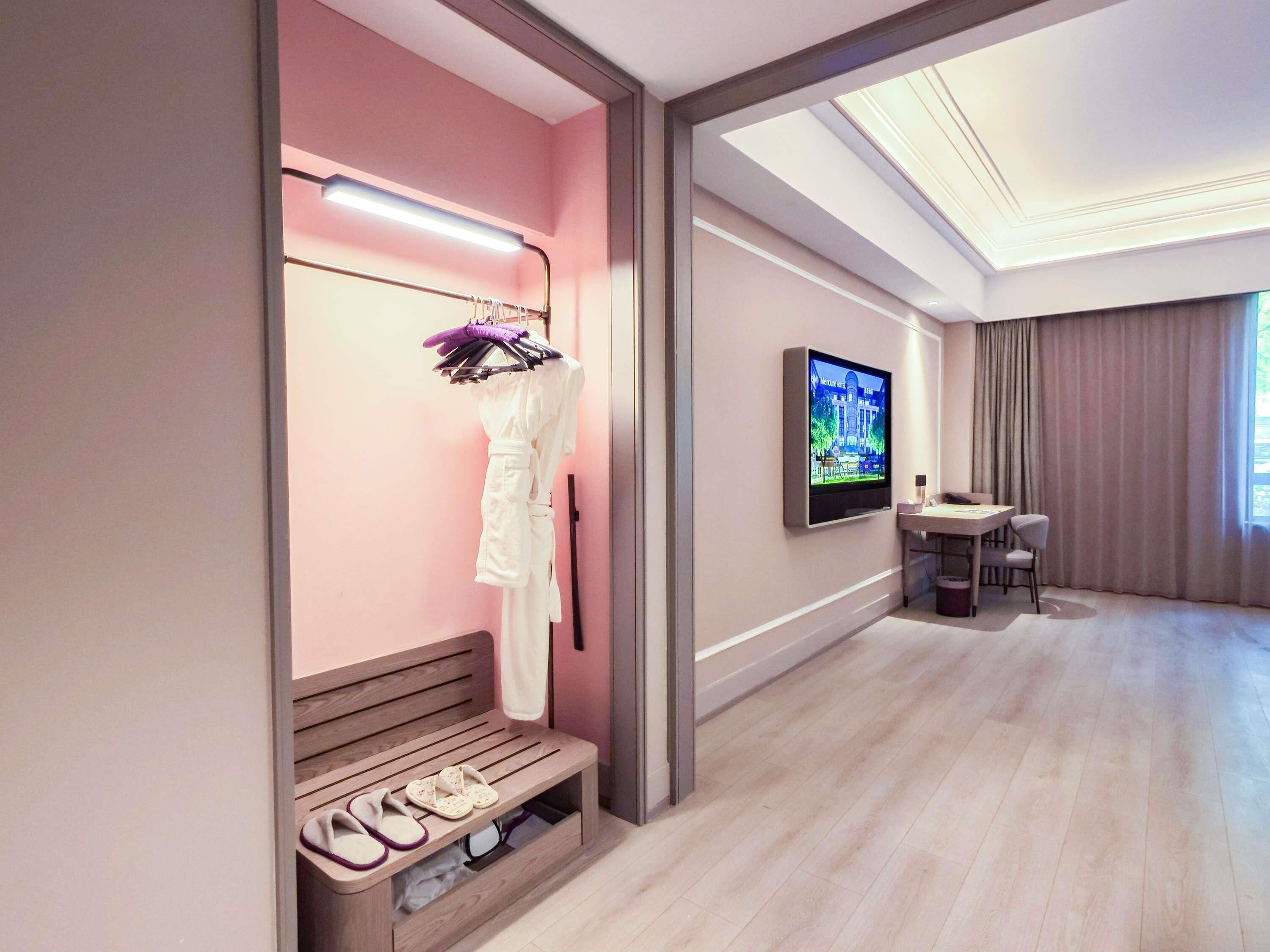 A hotel room with a vanity area, a television mounted on the wall, a seating area with a table and chairs, and a bench with slippers.