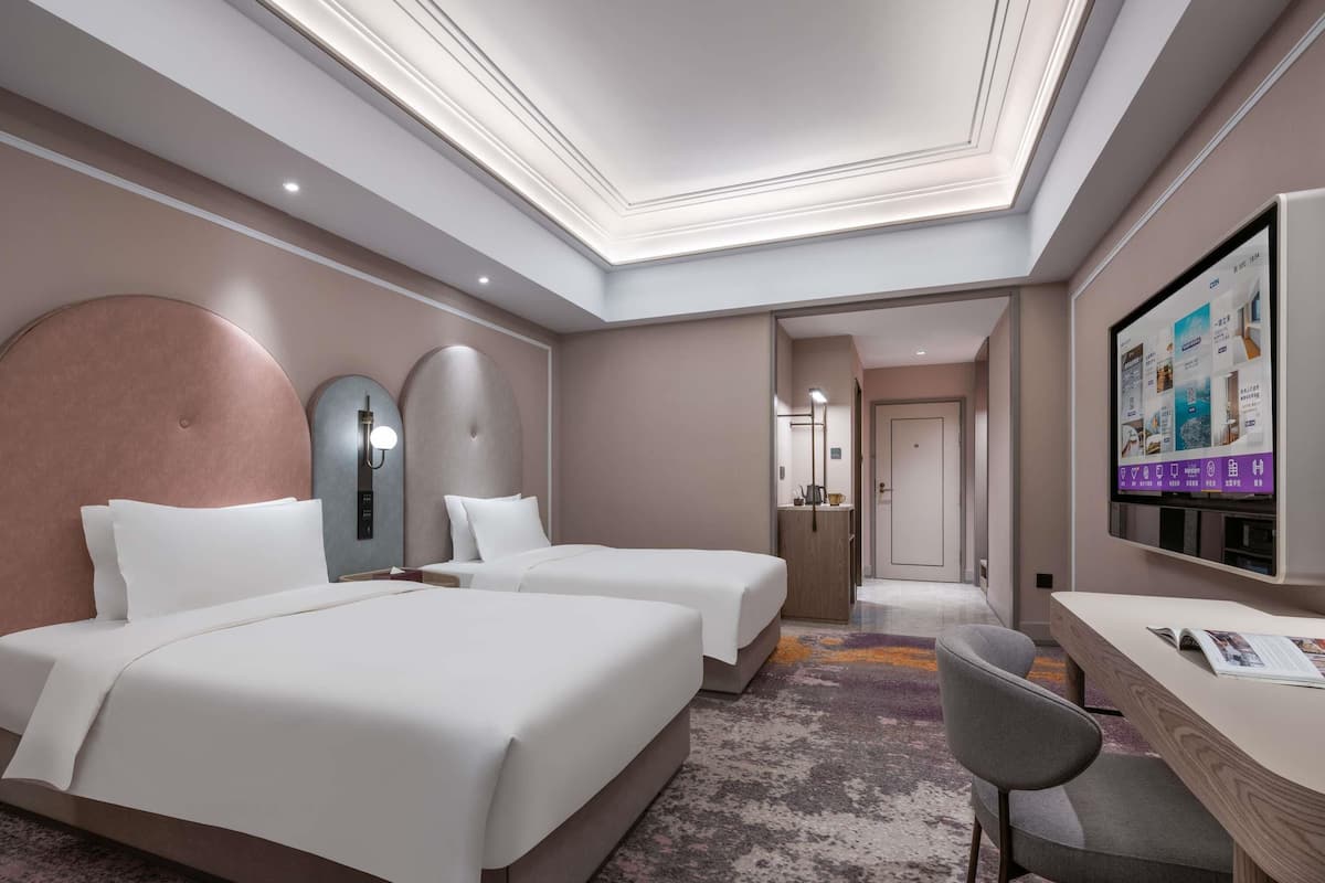 A hotel room with two beds, a desk, a chair, a TV, and a bathroom visible through an open door.