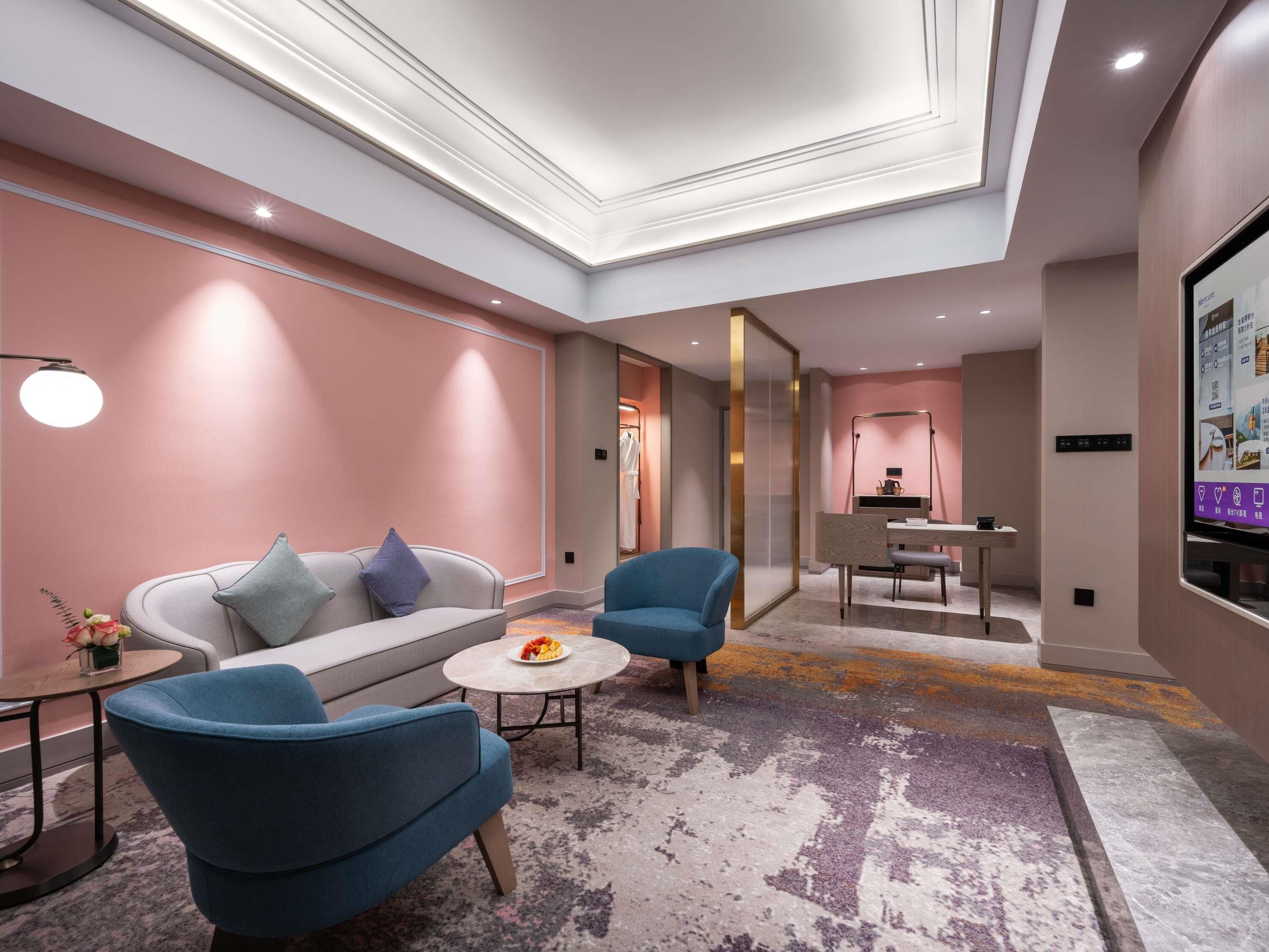 Ein modernes Hotelzimmer mit einer Sitzecke, zwei Sesseln, einem kleinen runden Tisch mit Früchten, einem Fernseher und einem Schreibtisch mit Stuhl.