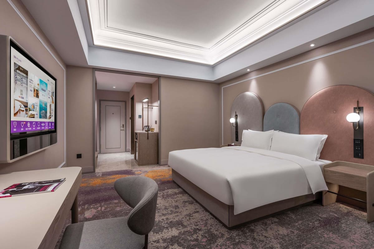 A modern hotel room with a large bed, a desk, a chair, and a television displaying room amenities.
