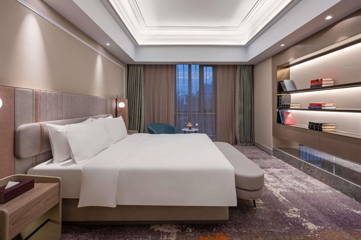 A hotel room with a large bed, bedside tables, a desk, a chair, a bookshelf, and a view of the outside.