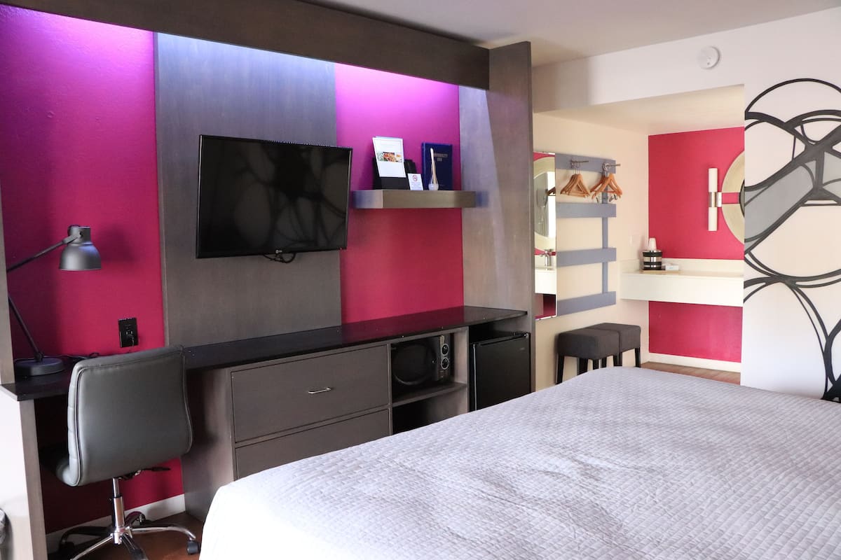 Une chambre d’hôtel moderne équipée d’un lit, d’un bureau avec une chaise, d’un téléviseur à écran plat fixé au mur et d’un petit coin cuisine.