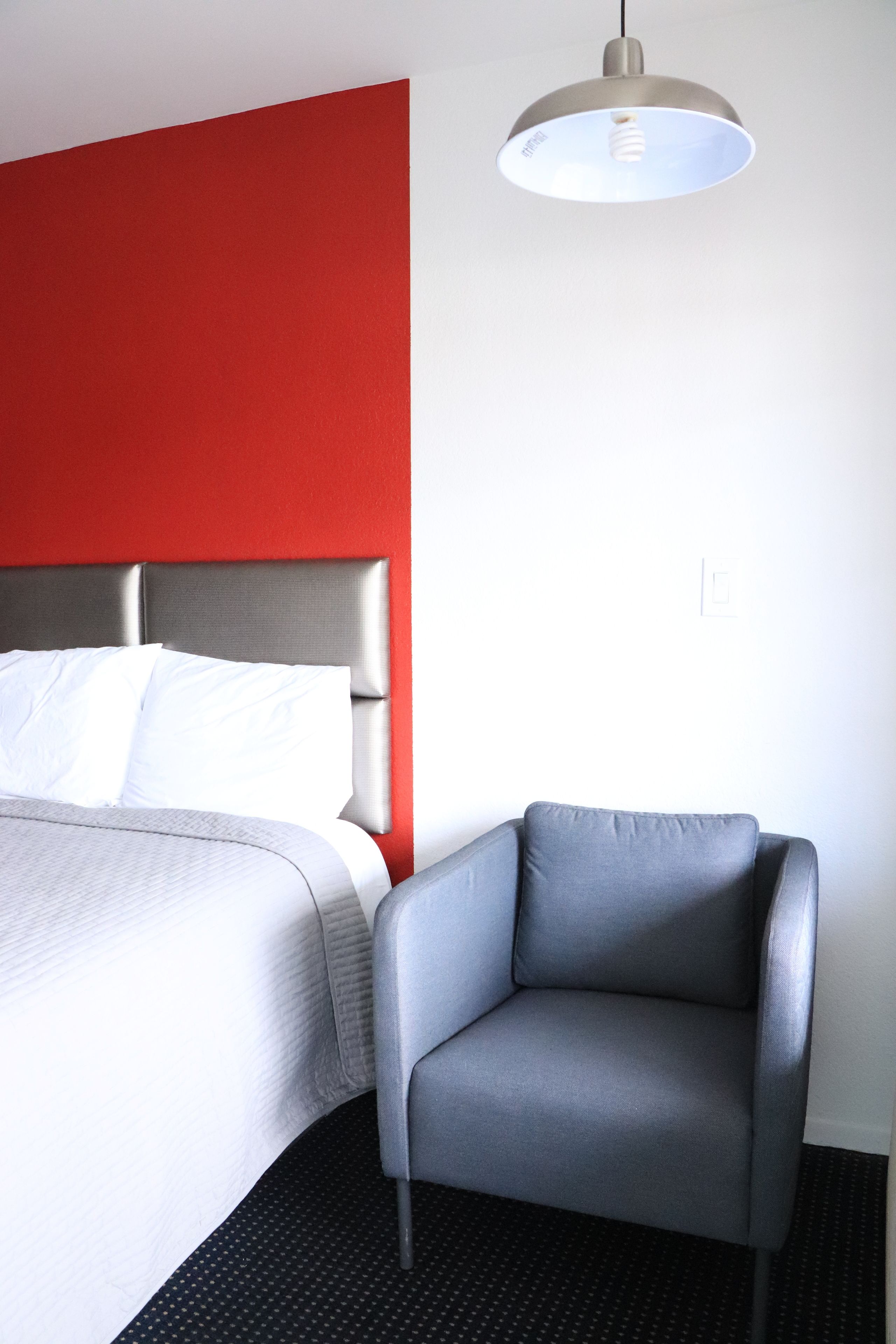 Habitación de hotel con cama, un sillón gris, pared de acento roja y lámpara en el techo.