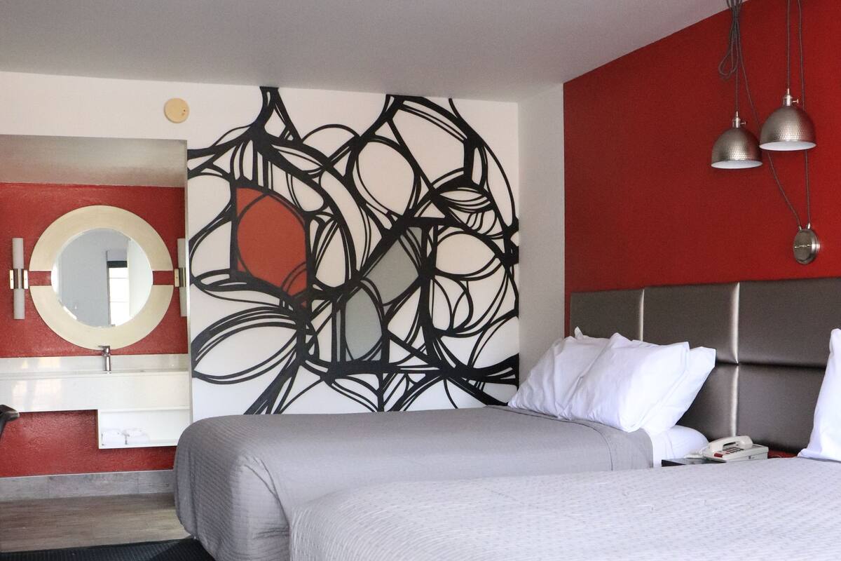 Une chambre d’hôtel moderne avec un grand lit, un miroir rond, un mur d’accent rouge, des œuvres d’art abstraites et un sol à motifs noir et blanc.