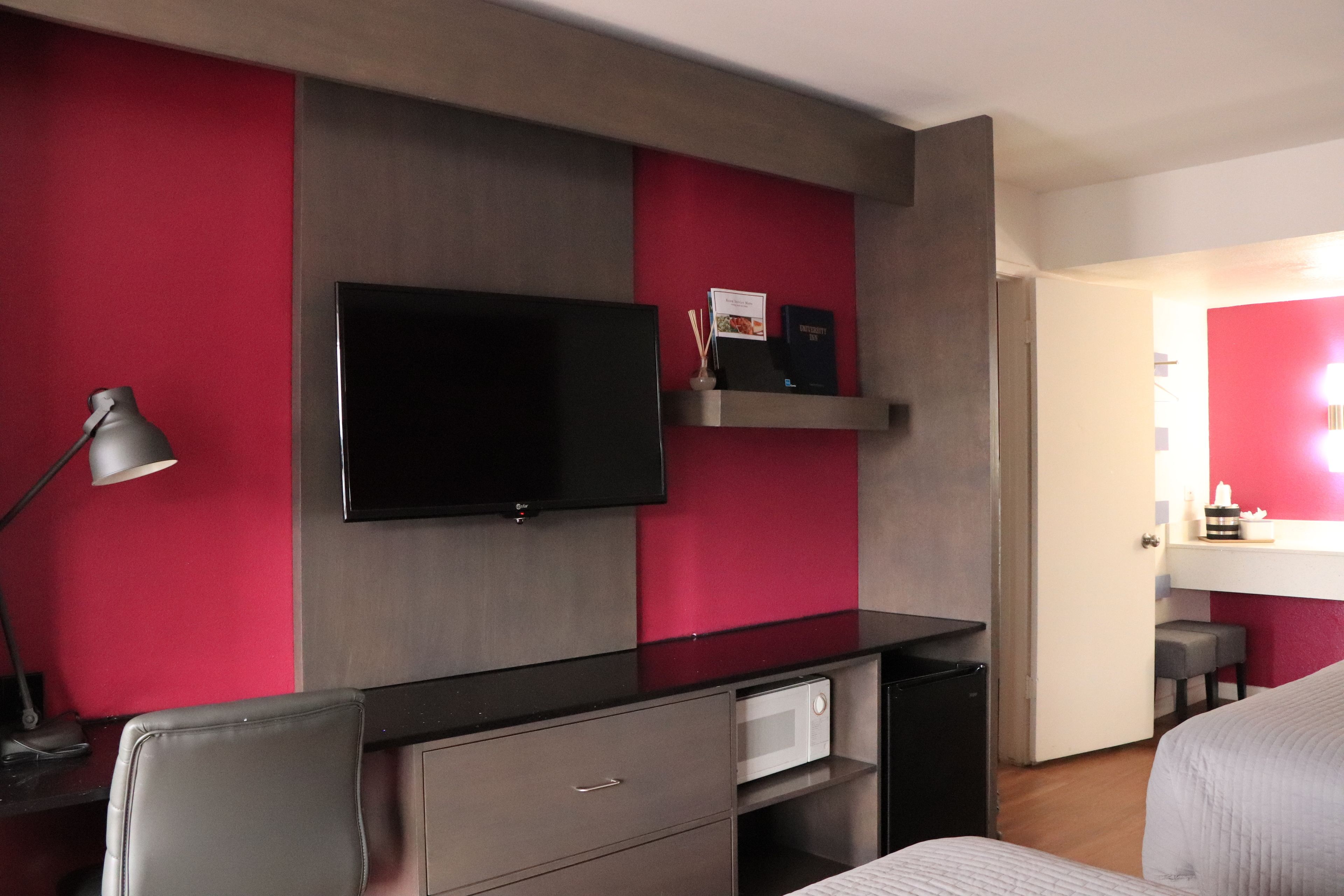 Una habitación de hotel moderna con una televisión de pantalla plana montada en la pared, un escritorio con lámpara, un microondas y un refrigerador pequeño.