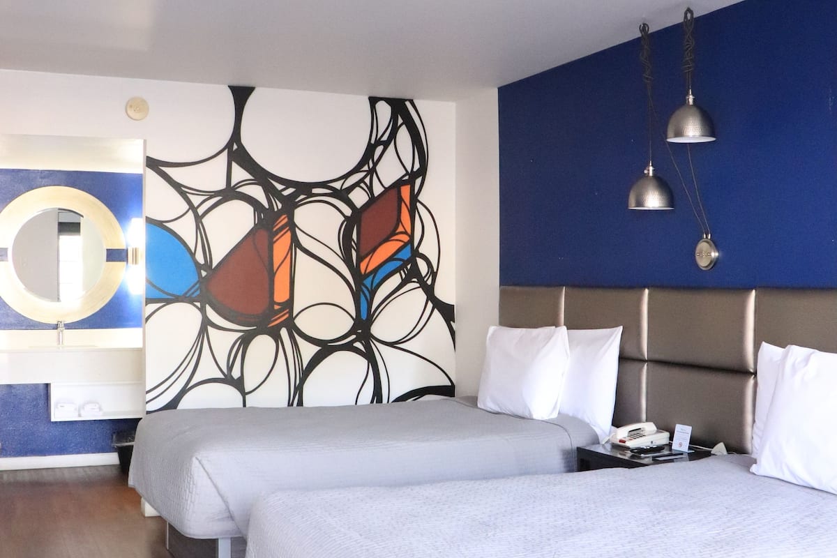 Une chambre d’hôtel avec deux lits, une œuvre d’art moderne et abstraite accrochée au mur, un mur d’accent bleu et un plancher en bois.