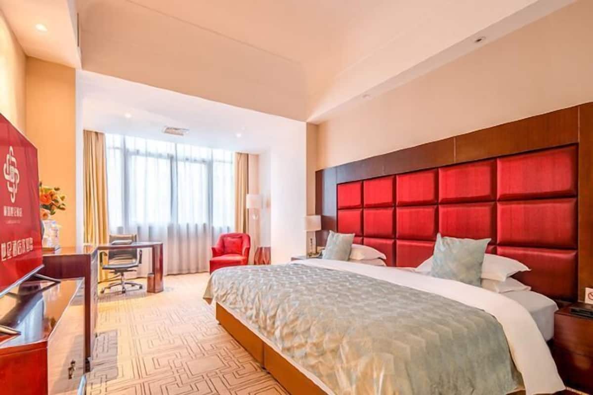 A hotel room with a large bed, a red headboard, a desk with a computer, and a window with curtains.