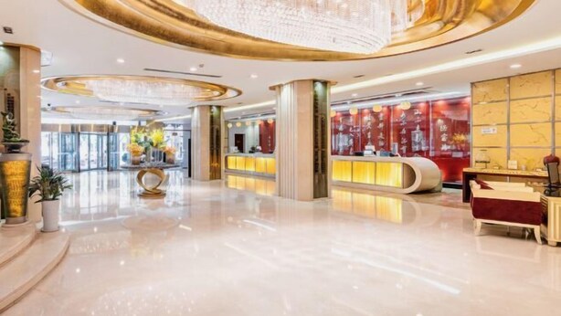 Eine großzügige Lobby mit spiegelndem Boden, einem zentralen Kronleuchter und einer Rezeption mit rotem Hintergrund.