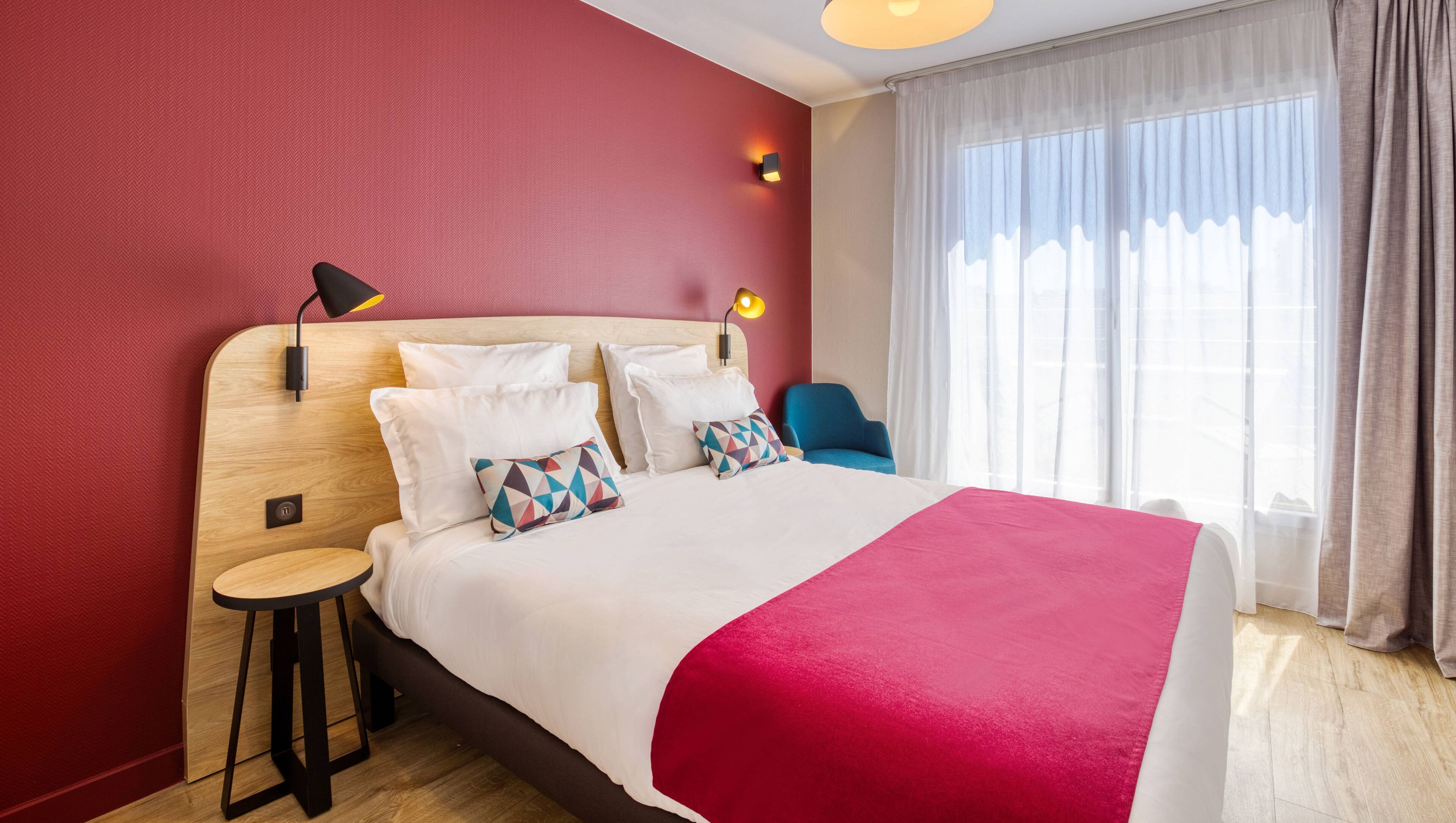 A hotel room with a large bed, a bedside table, a chair, a lamp, and a window with curtains.