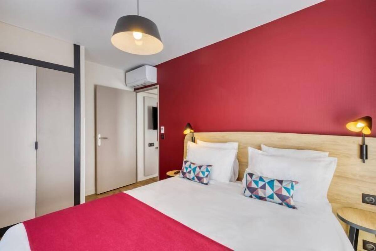Una habitación de hotel con una cama, una pared de acento roja, una lámpara en el techo y una mesita de noche.