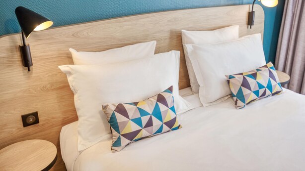 Habitación de hotel con cabecera de madera, dos almohadas blancas, una almohada geométrica colorida, una mesita redonda y una lámpara de pared.