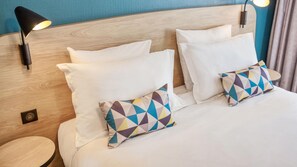 A hotel room with a wooden headboard, two white pillows, a colorful geometric pillow, a small round table, and a wall-mounted lamp. - Appart'City Confort Paris Clichy Mairie (Clichy)