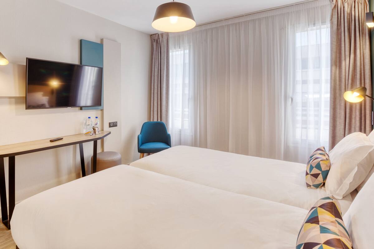 Habitación de hotel con dos camas, un escritorio con televisor, una silla y una ventana con cortinas.