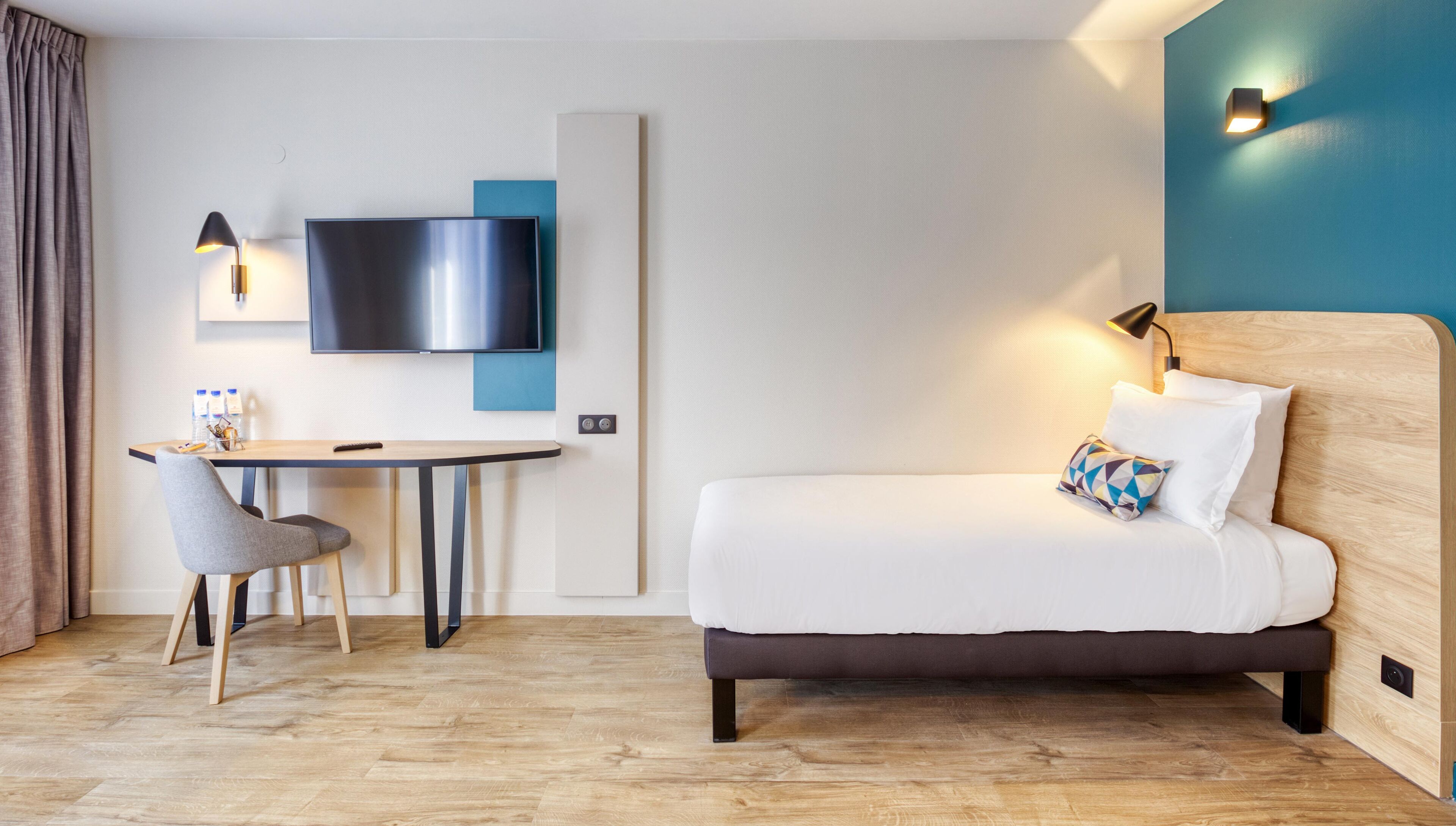 Une chambre d’hôtel équipée d’un lit, d’un bureau, d’une chaise, d’une télévision et d’une lampe fixée au mur.