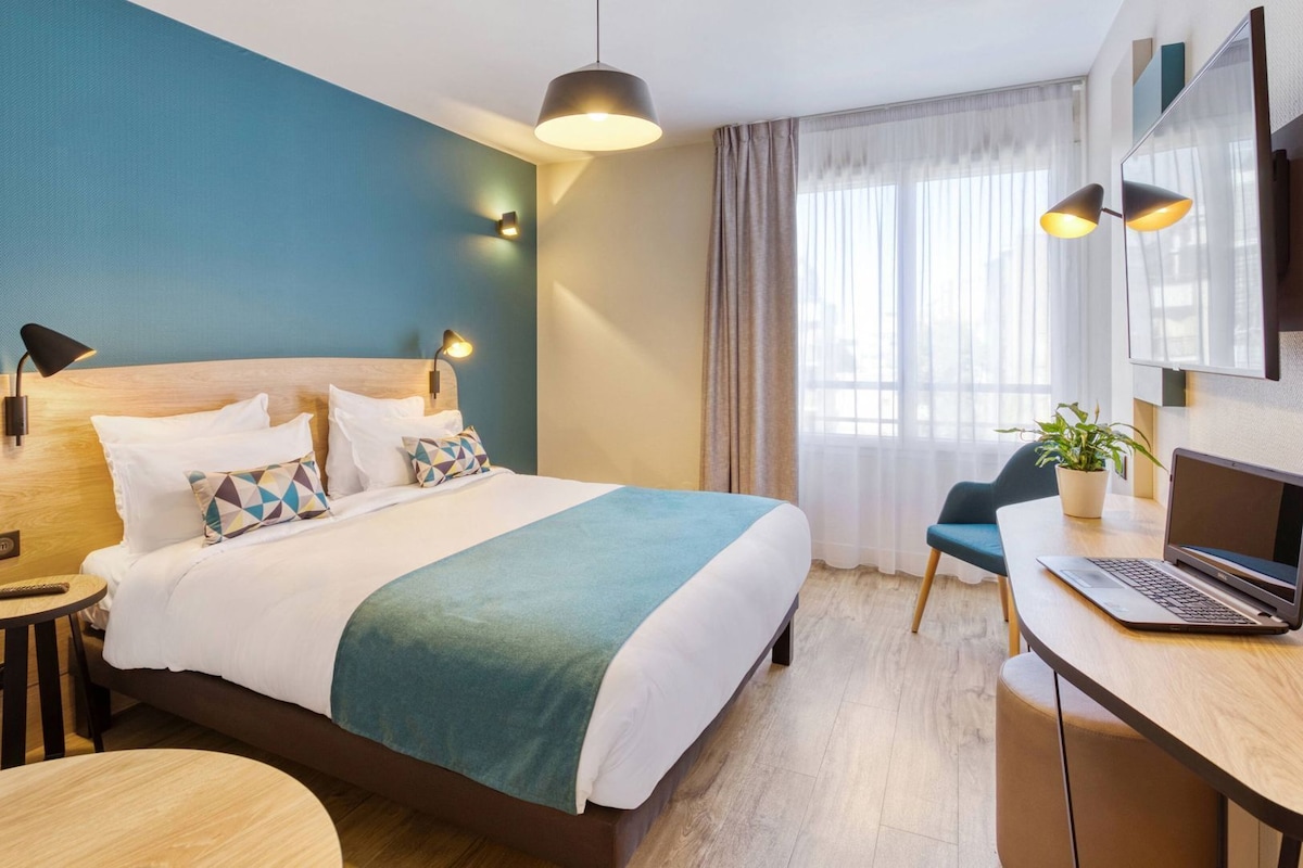 Habitación de hotel con cama, mesita de noche, lámpara, escritorio con una computadora portátil, silla y planta en maceta.