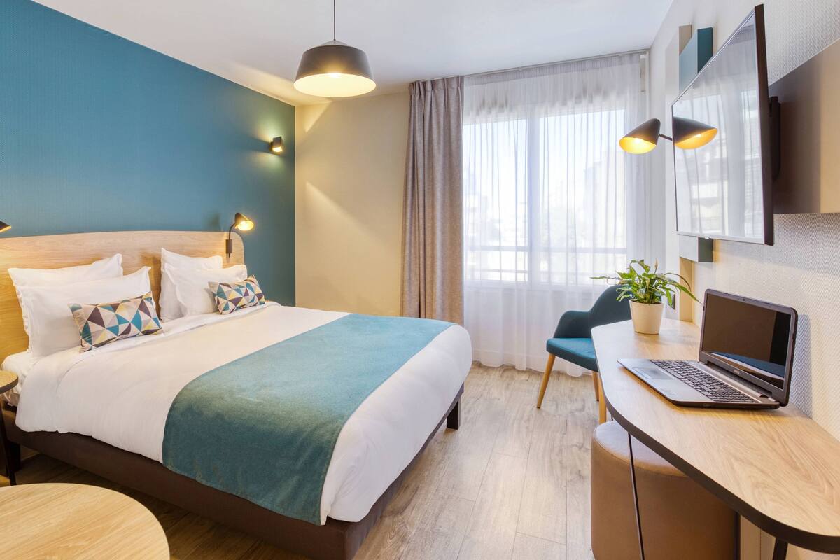 Una habitación de hotel moderna con una cama de madera, un escritorio con una computadora portátil, una mesita redonda y una planta en maceta.
