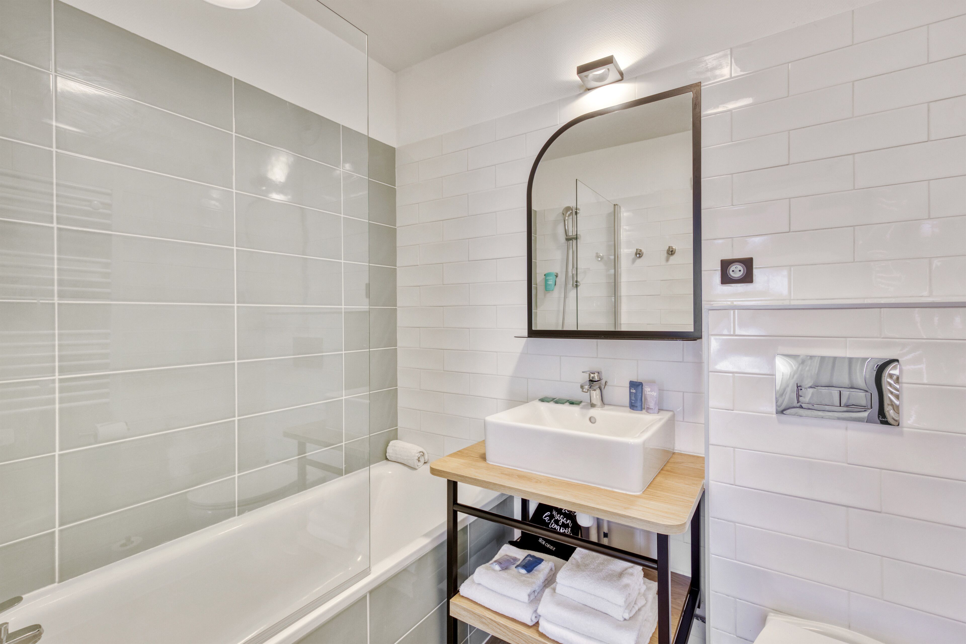A modern bathroom with a white bathtub, a sink with a mirror above it, and a shelf with towels.