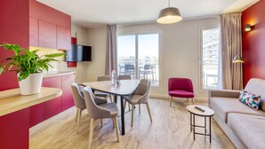 Una habitación de hotel moderna con zona de comedor, sofá, televisor y un amplio ventanal con vistas.