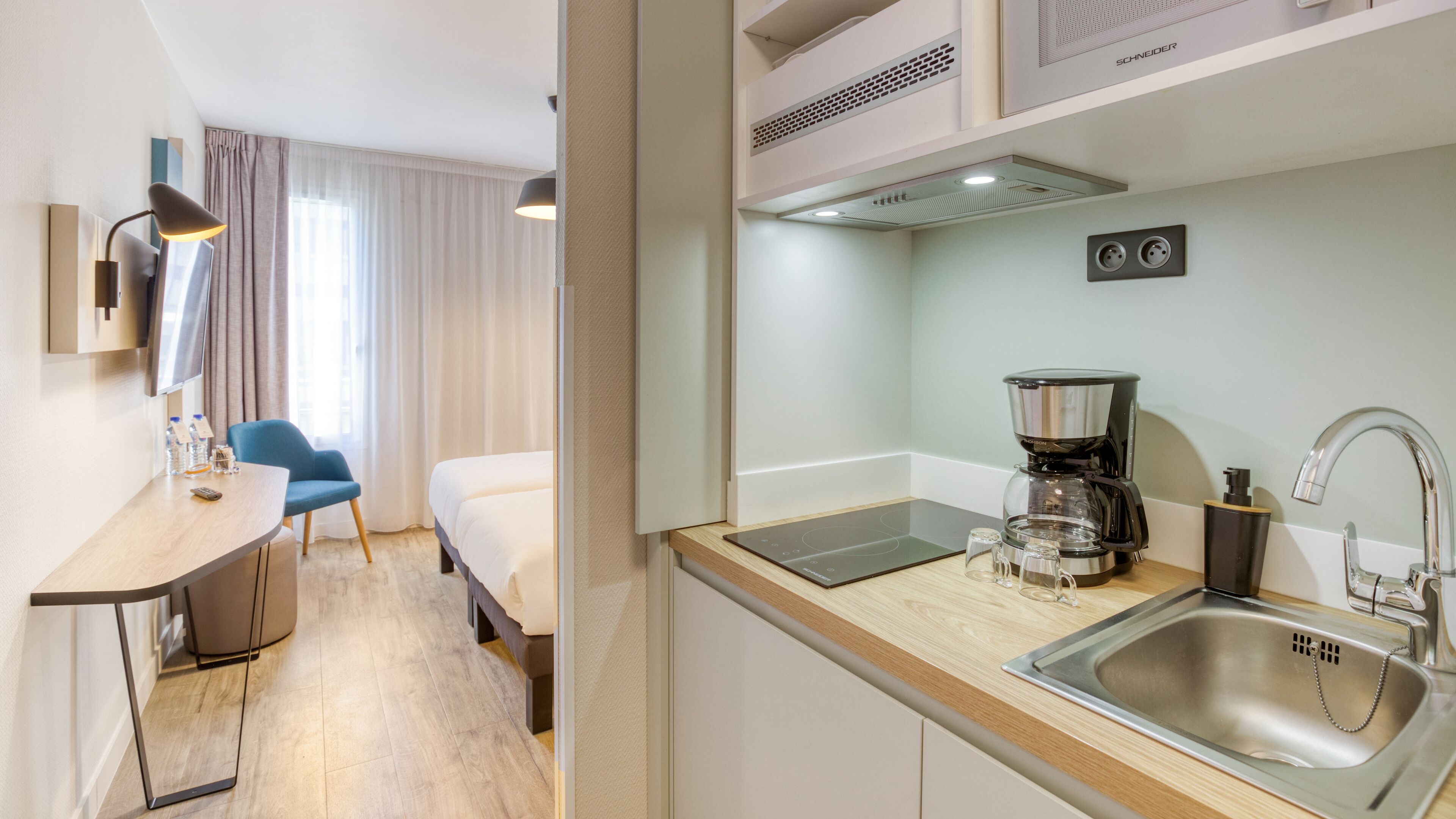 A compact hotel room with a kitchenette, a bed, a desk with a chair, and a window with curtains.