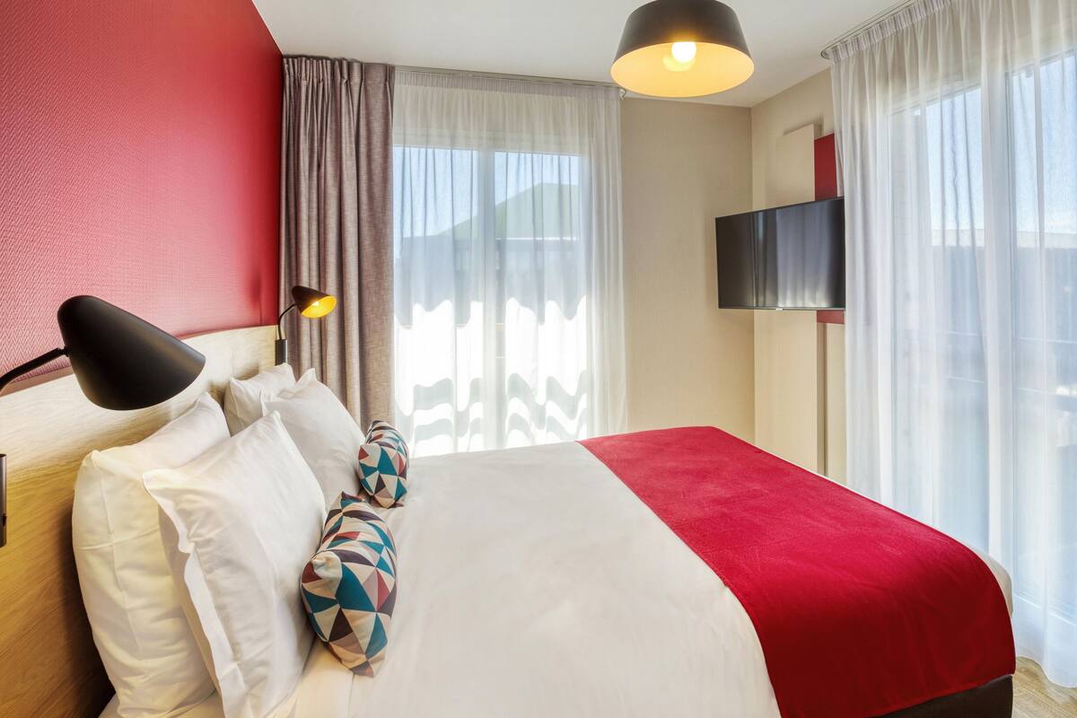 Una habitación de hotel con cama, televisión y ventana con cortinas.