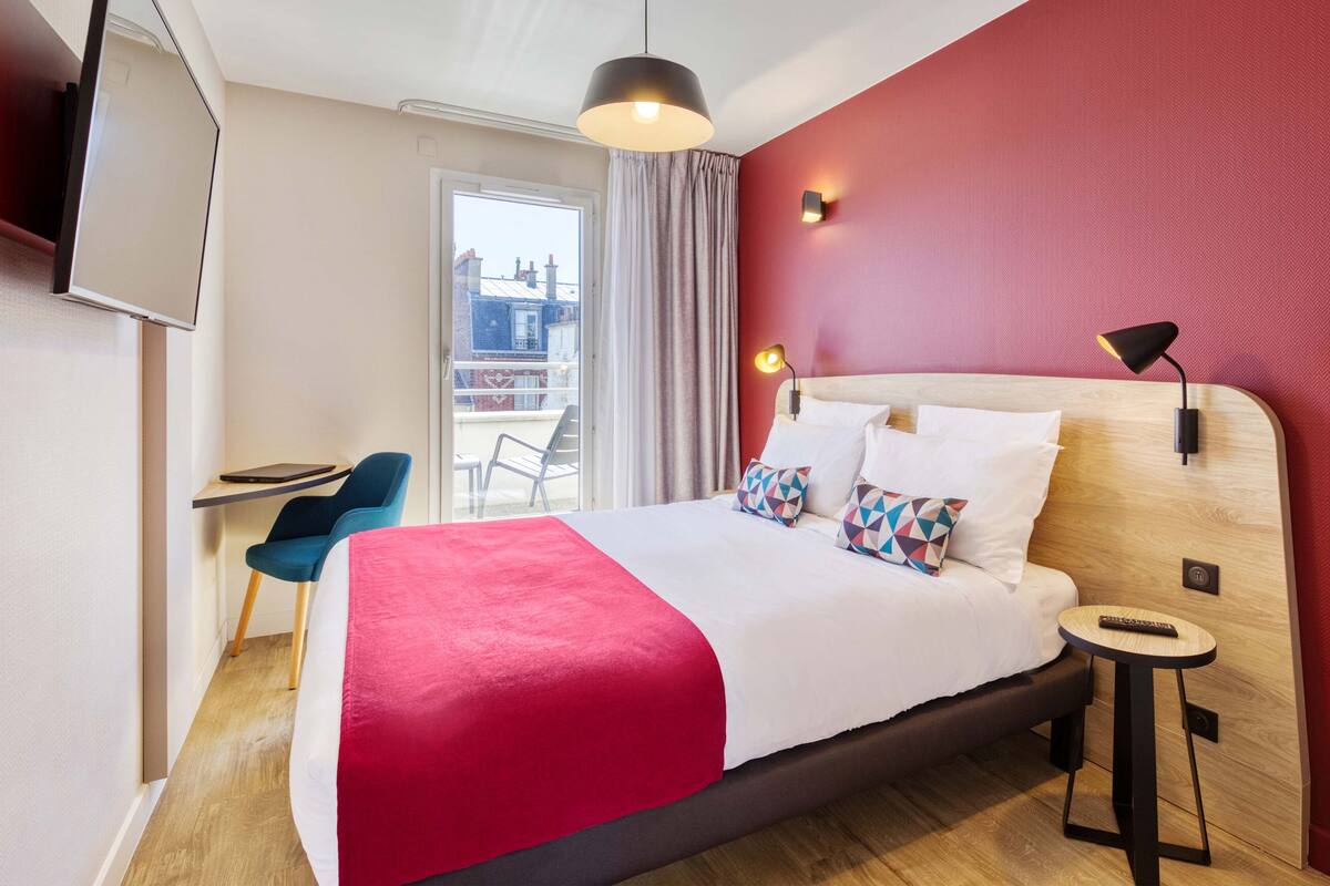Una habitación de hotel con cama, escritorio, silla, ventana con cortinas y vistas a edificios.
