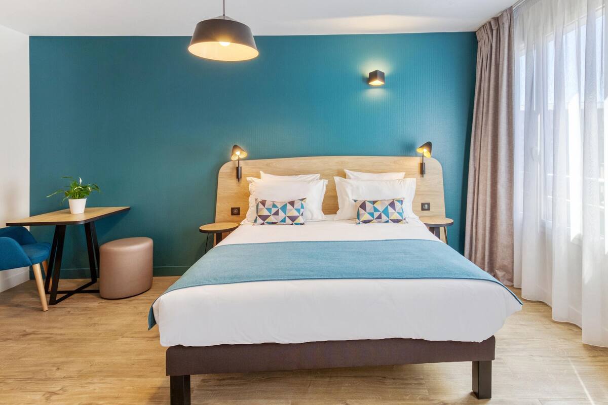 Una habitación de hotel moderna con una cama grande, un cabecero de madera, dos mesitas de noche, una pequeña mesa redonda, una silla azul, una planta en maceta y vistas al exterior a través de cortinas translúcidas.