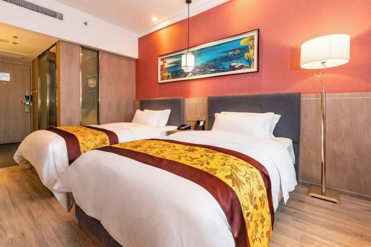 A hotel room with two beds, a painting on the wall, a floor lamp, and a door with a glass panel.