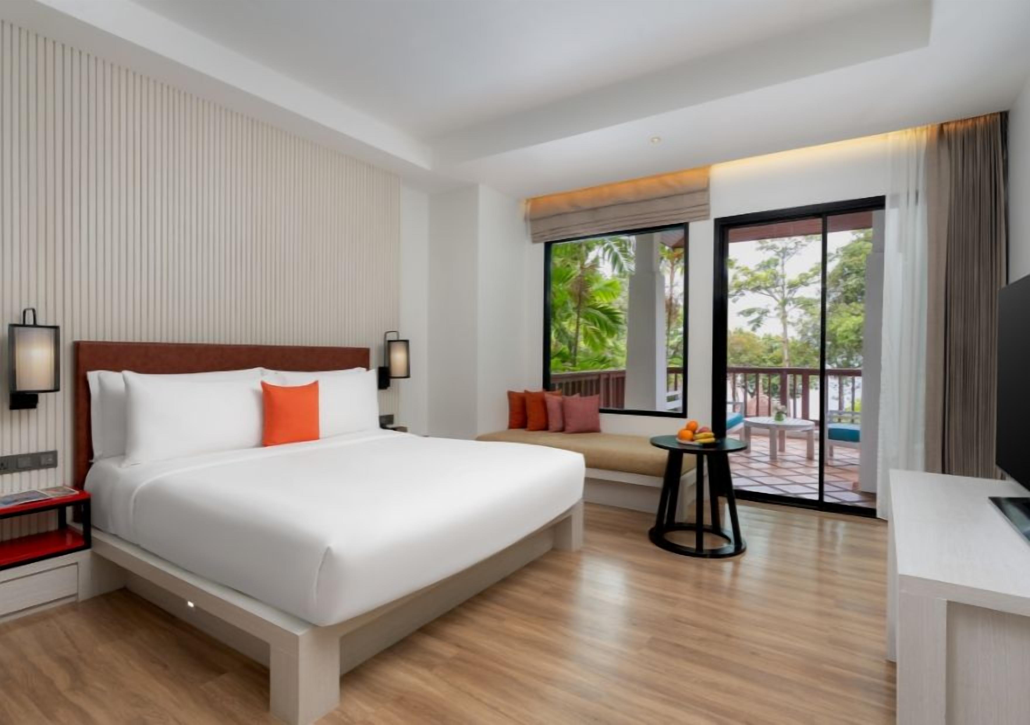 A hotel room with a large bed, a sofa, a TV, and a balcony with a view of trees.