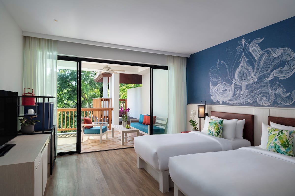 Habitación de hotel con dos camas, un escritorio, televisión y balcón con mobiliario exterior.