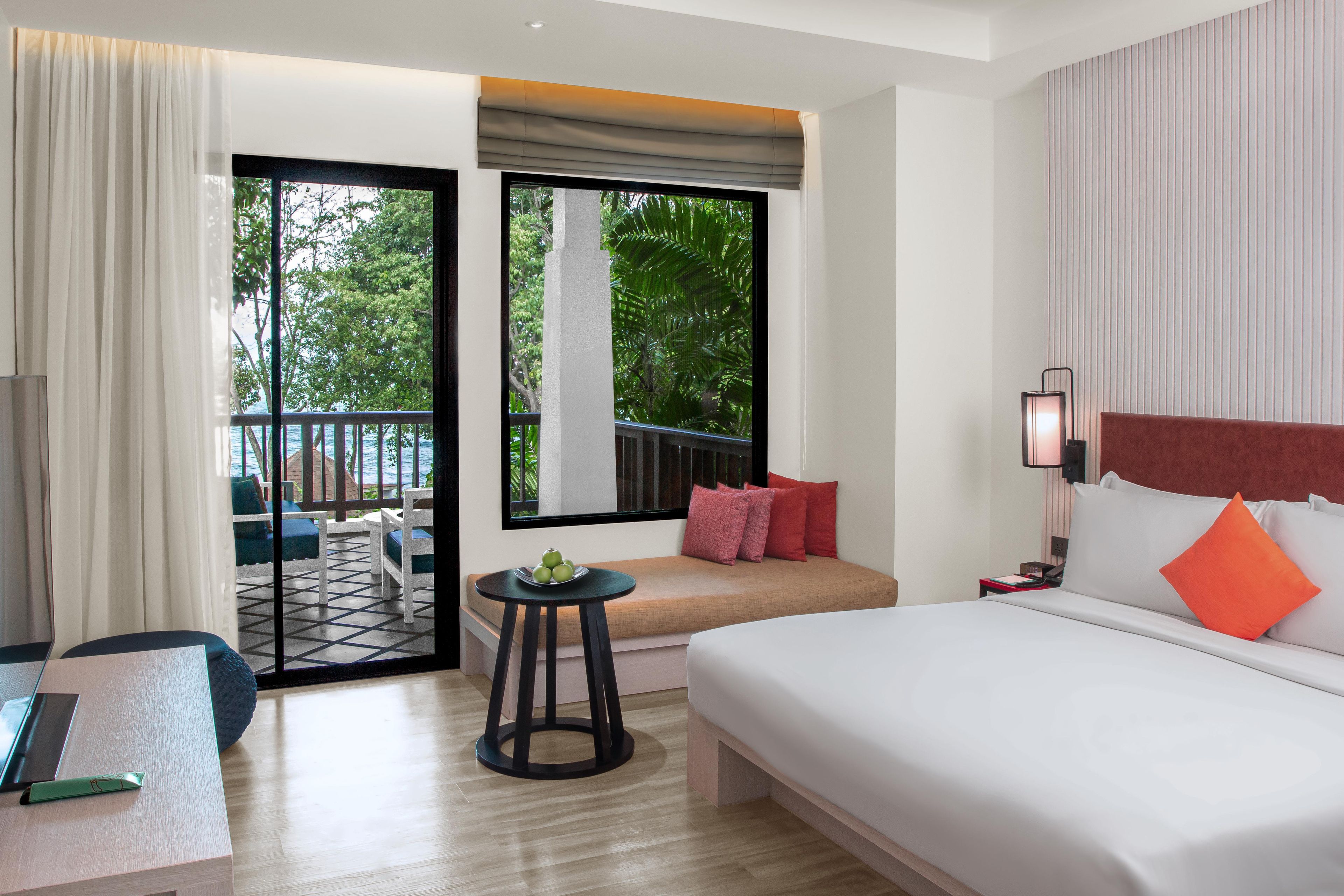 A hotel room with a bed, a small table, a chair, a sofa, and a balcony with a view of greenery.