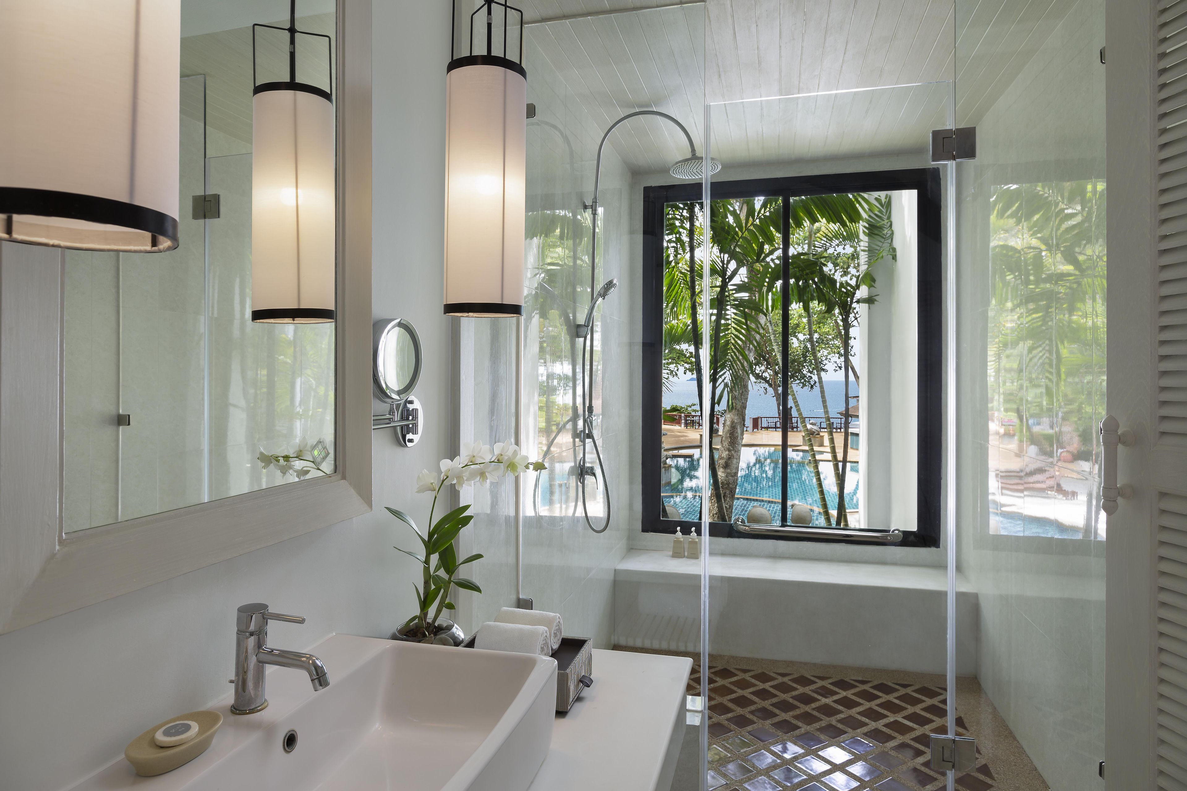 Une salle de bain moderne avec un grand miroir, deux lavabos, une douche et une fenêtre donnant sur une piscine et des palmiers.
