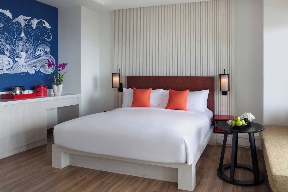 Een hotelkamer met een groot bed, een nachtkastje met een schaal fruit, een rode stoel en een decoratief wandobject.