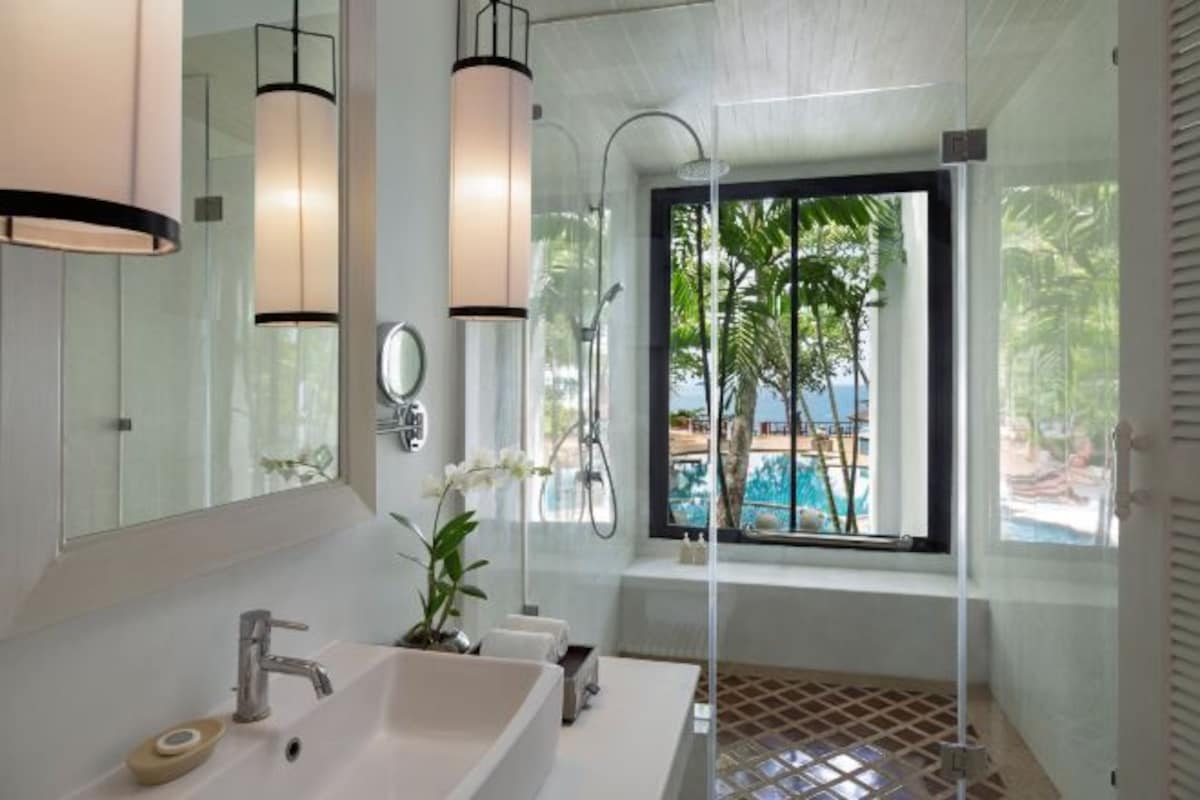 Un baño moderno con un espejo grande, un lavamanos blanco, ducha y ventana con vista a una piscina.