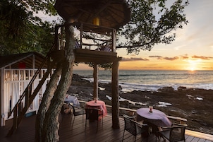 Una terraza de madera con mobiliario exterior con vistas a una costa rocosa al atardecer.