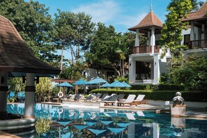 A pool area with lounge chairs and umbrellas, surrounded by lush greenery and a building with a distinctive roof.