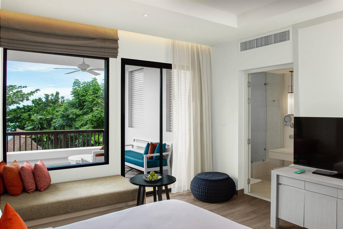 Habitación de hotel con balcón, cama, televisor, una pequeña mesa con un cuenco de fruta y una zona de estar con sofá y sillas.
