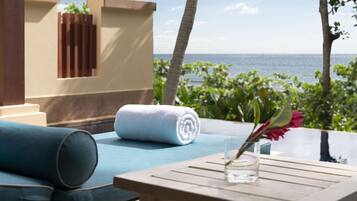 A sunlit outdoor seating area with a view of the ocean, a rolled white towel, and a glass vase with a flower.