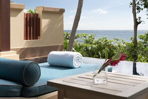 A sunlit outdoor seating area with a view of the ocean, a rolled white towel, and a glass vase with a flower.